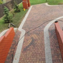 ProExtreme Tomasz Szempliński - Ułożona kostka brukowa w odcieniach brązu z jasnoszarymi obrzeżami, widok z góry na posesję z trawnikiem, drzewami i jeziorem w tle, elementem grilla z cegły i żółtym wiadrem.