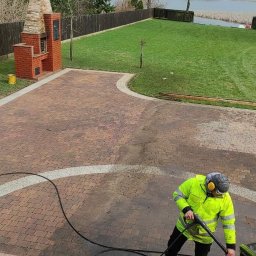 ProExtreme Tomasz Szempliński - Pracownik w odblaskowej kurtce czyści kostkę brukową na posesji z grillem murowanym i widokiem na jezioro przy użyciu myjki ciśnieniowej, widoczny kontrast czystej i brudnej powierzchni.