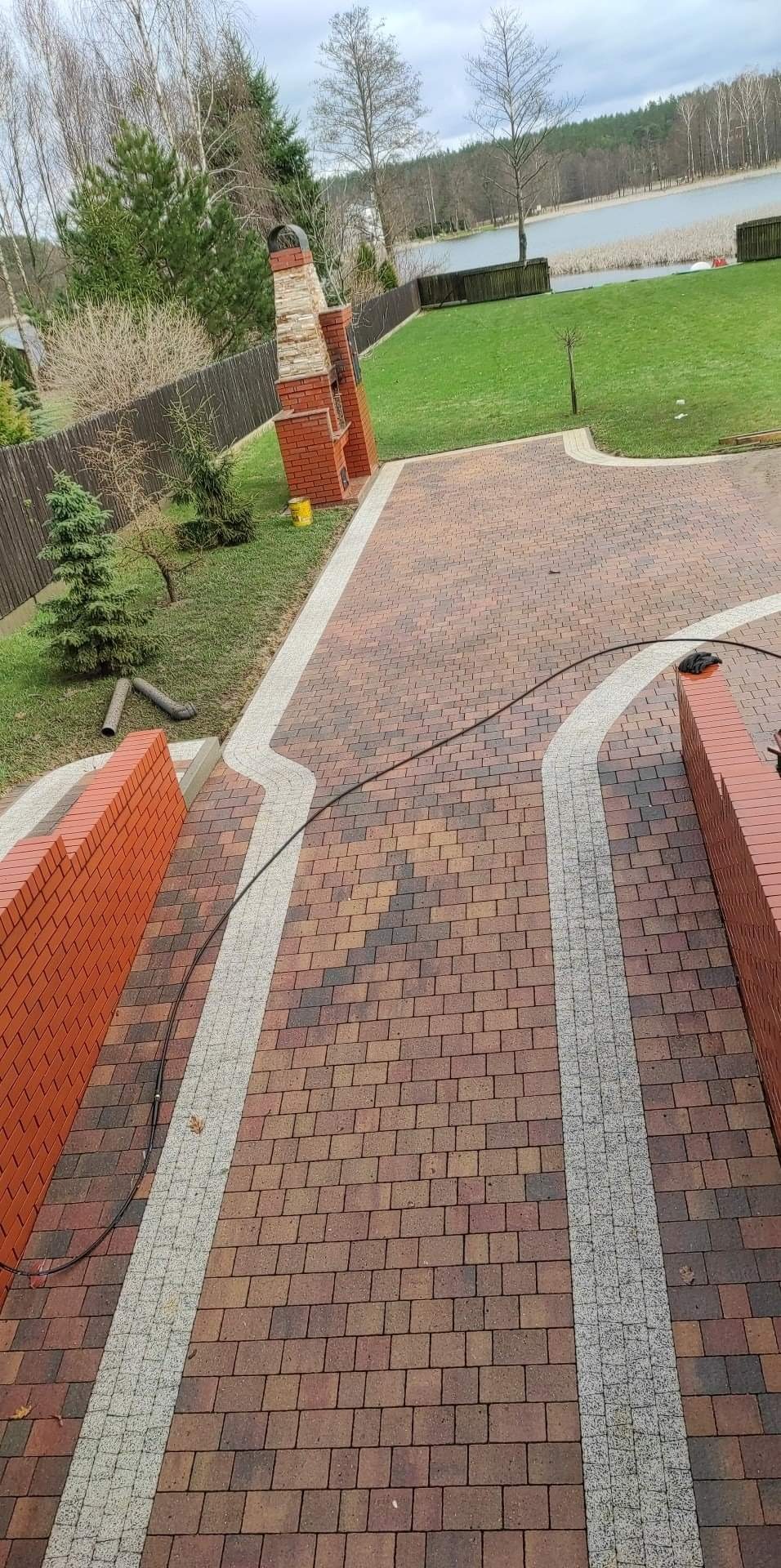 Ułożona kostka brukowa w odcieniach brązu z jasnoszarymi obrzeżami, widok z góry na posesję z trawnikiem, drzewami i jeziorem w tle, elementem grilla z cegły i żółtym wiadrem.