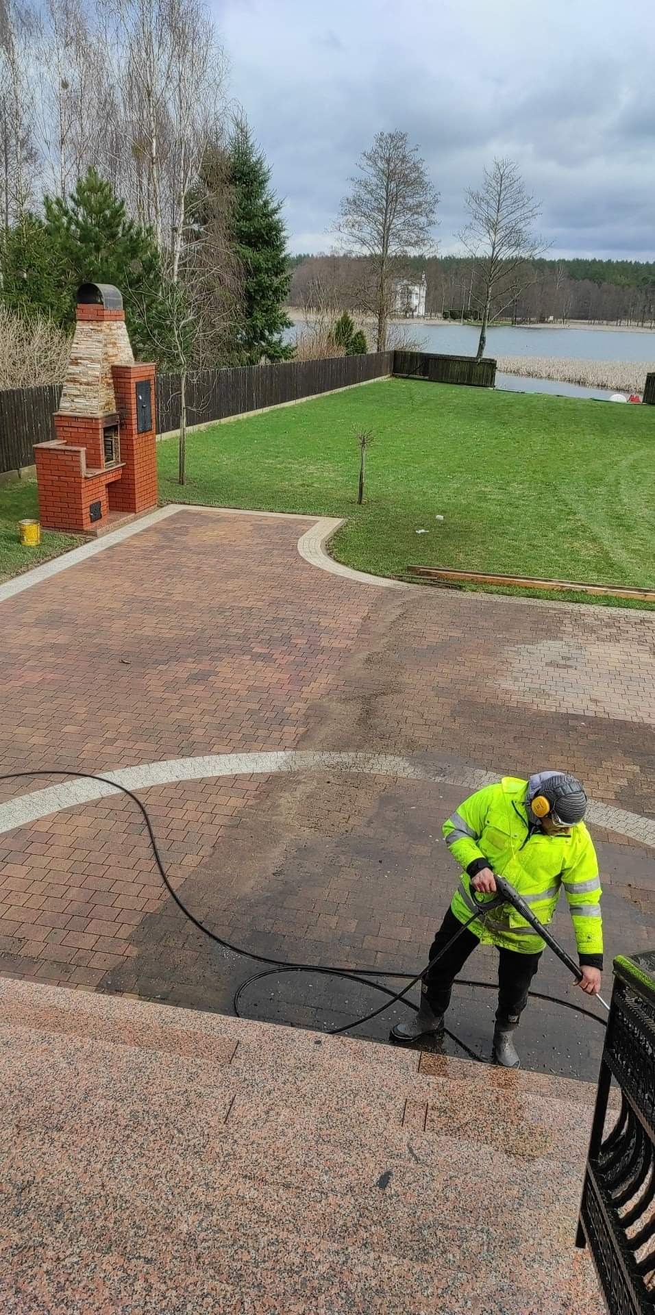 Pracownik w odblaskowej kurtce czyści kostkę brukową na posesji z grillem murowanym i widokiem na jezioro przy użyciu myjki ciśnieniowej, widoczny kontrast czystej i brudnej powierzchni.