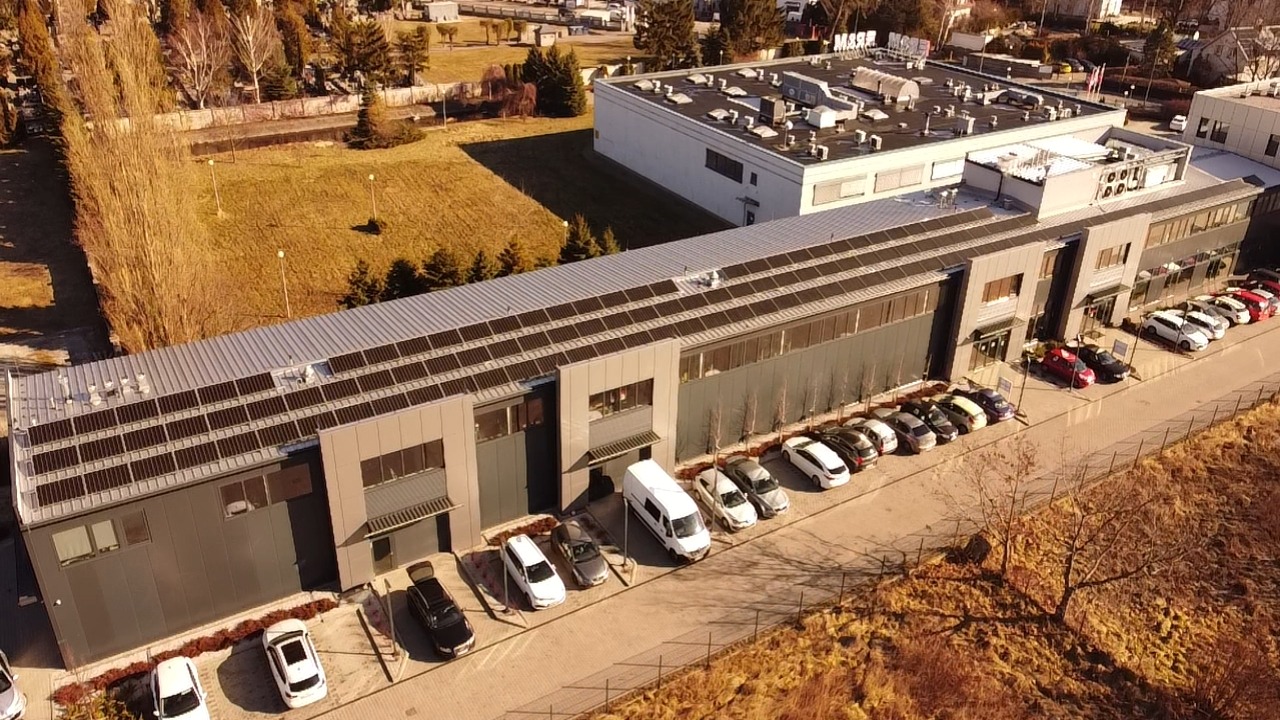 Widok z lotu ptaka na budynek przemysłowy z panelami słonecznymi na dachu, zaparkowanymi samochodami i przyległym terenem zielonym.