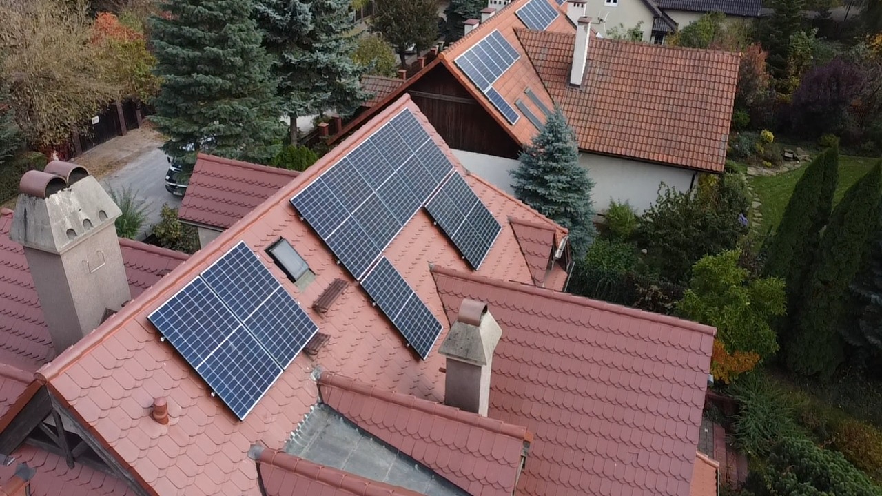 Widok z góry na dachy domów z zainstalowanymi panelami fotowoltaicznymi; czerwona dachówka, ciemne panele, kominy, drzewa w tle.