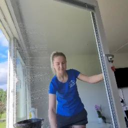 Kobieta w niebieskiej koszulce z logo firmy czyści okno za pomocą ściągaczki, stojąc na składanym stołku; widoczny czarny wiadro z wodą i płynem do mycia okien, oraz widok na zewnątrz przez umyte...