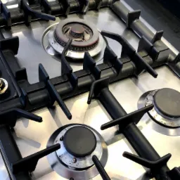 Szczegółowe ujęcie płyty gazowej ze stali nierdzewnej z czarnymi żeliwnymi rusztami, widoczne zabrudzenia i ślady użytkowania na palnikach.