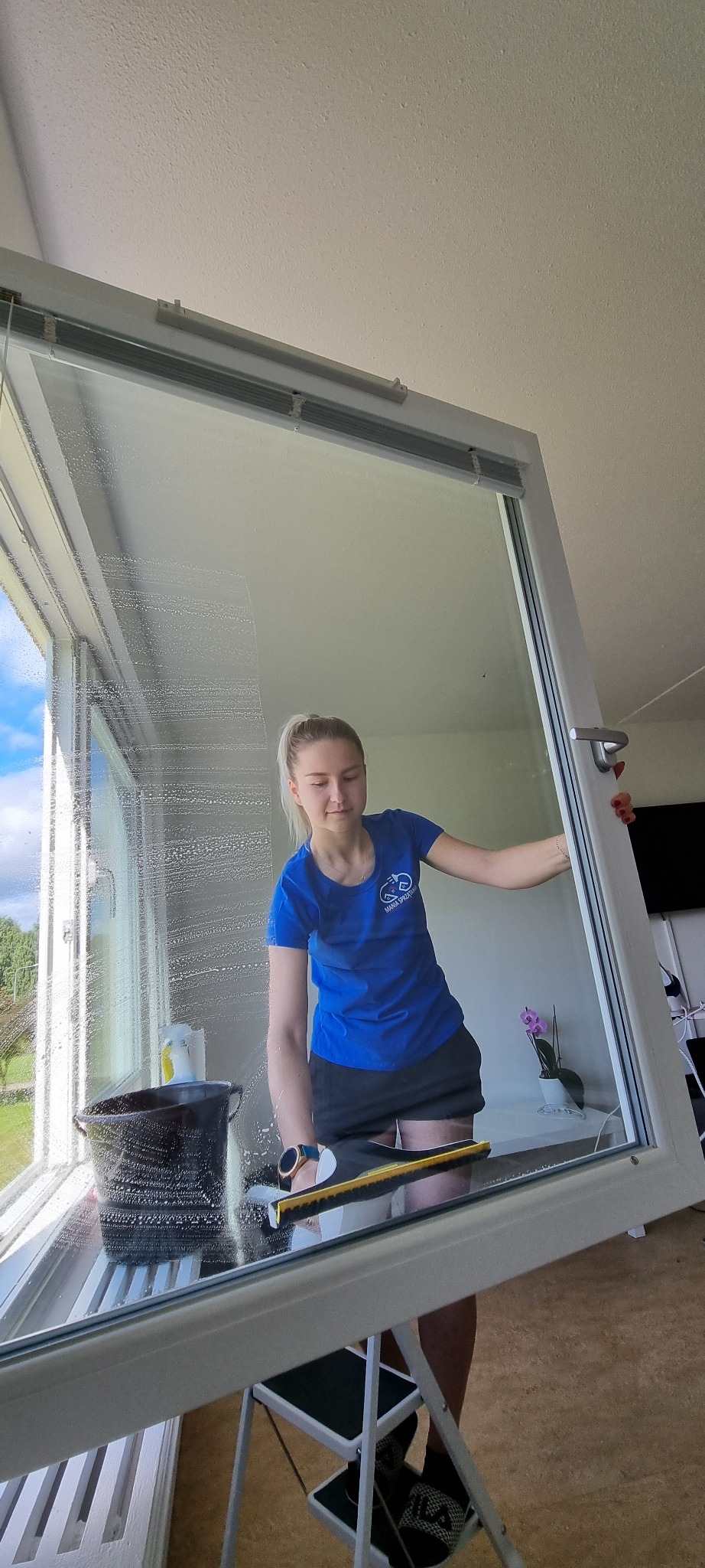 Kobieta w niebieskiej koszulce z logo firmy czyści okno za pomocą ściągaczki, stojąc na składanym stołku; widoczny czarny wiadro z wodą i płynem do mycia okien, oraz widok na zewnątrz przez umyte...