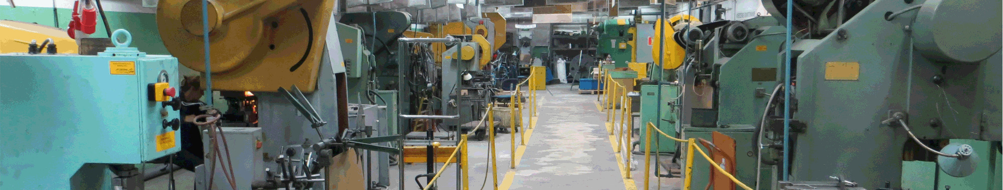 Wnętrze hali produkcyjnej z rzędem obrabiarek CNC, widok wzdłuż korytarza wyznaczonego żółtymi barierkami, maszyny w kolorach zielonym, żółtym i turkusowym, oświetlenie przemysłowe.