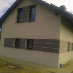 Nowo otynkowany dom jednorodzinny z szarymi, poziomymi pasami dekoracyjnymi wokół okien, widoczne rusztowanie i teren budowy.