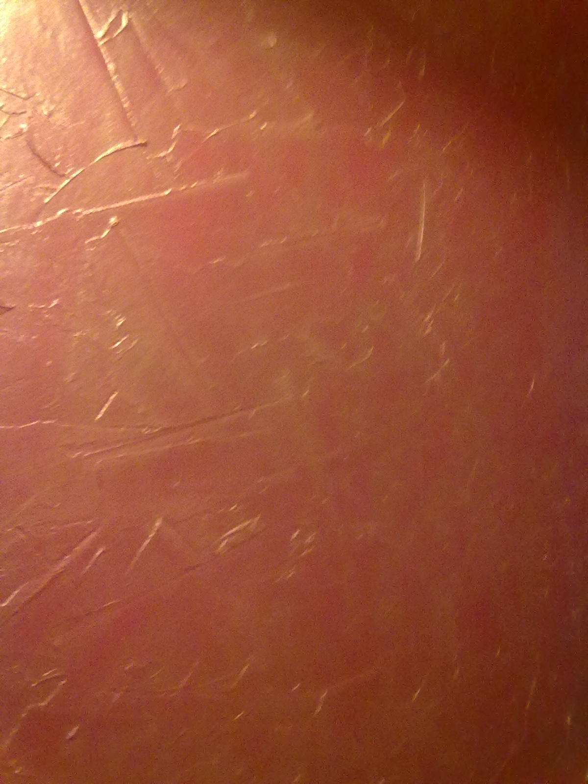 Nierówna, różowo-złota powierzchnia ściany z widocznymi smugami i niedoskonałościami gładzi, oświetlona ciepłym światłem.