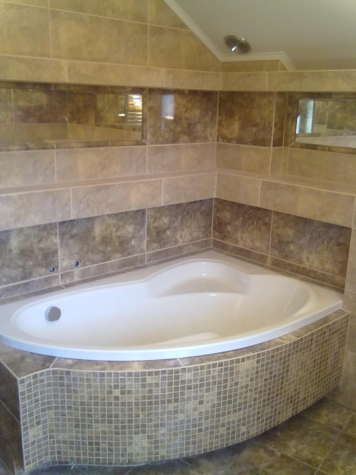 Narożna wanna obudowana mozaiką w łazience z beżowo-brązowymi płytkami, z dwoma wnękami z lustrami w ścianie.