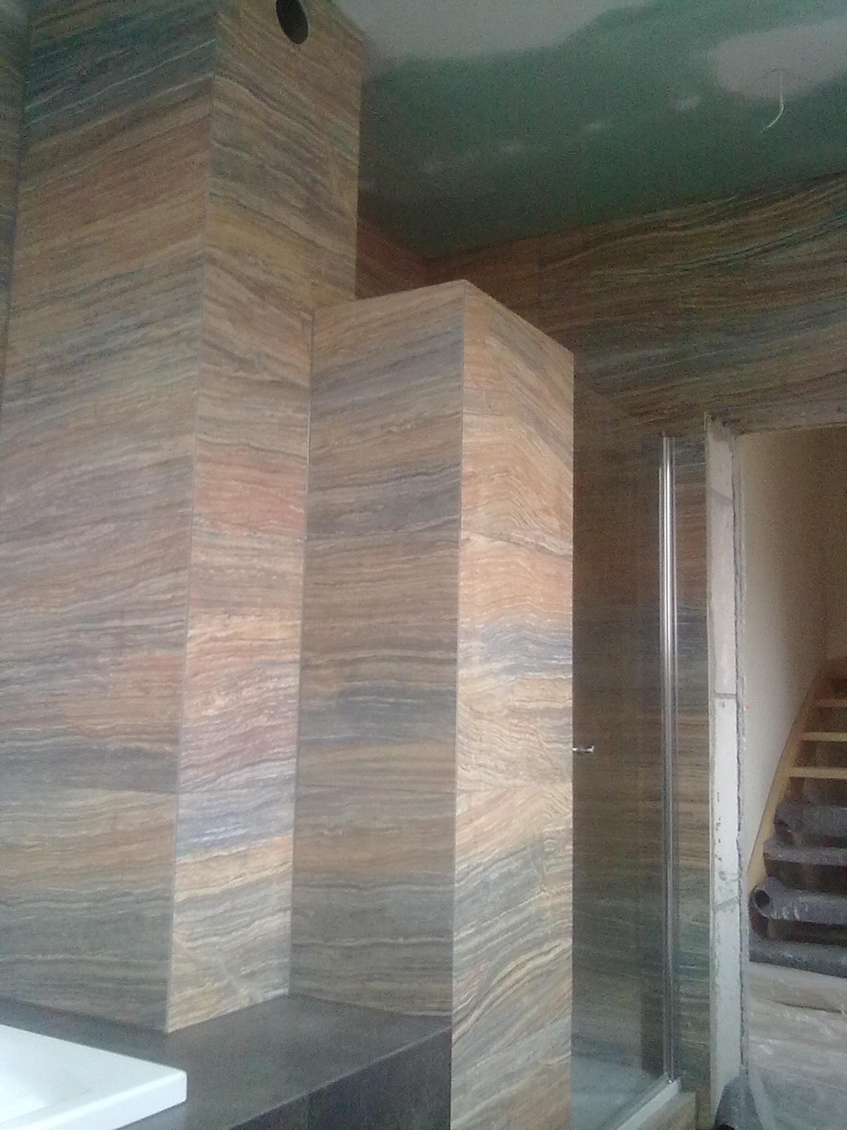 Ściany w łazience wyłożone płytkami imitującymi warstwową skałę, widoczna kabina prysznicowa i fragment schodów w tle, sufit w stanie surowym.