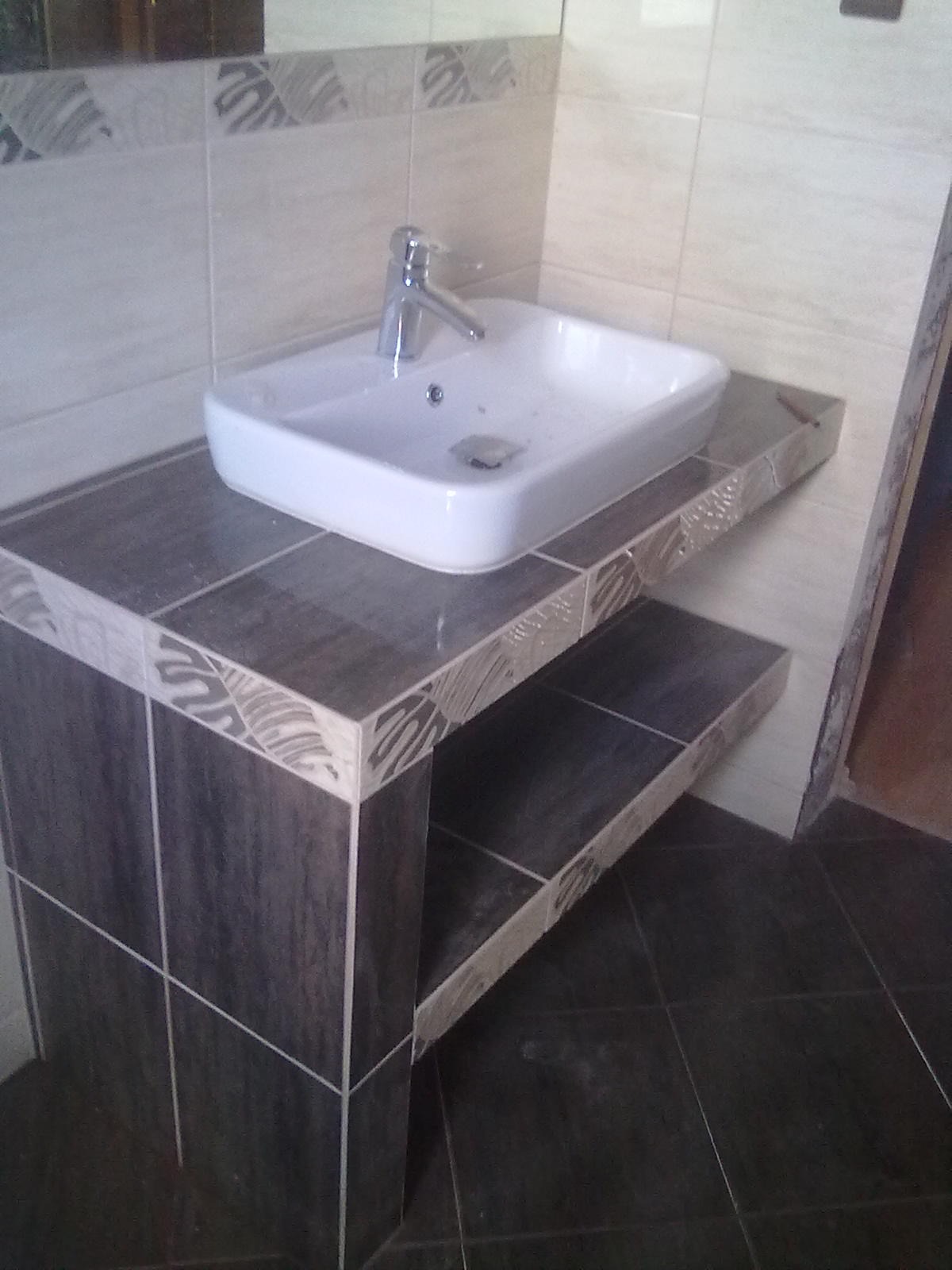 Biała, prostokątna umywalka nablatowa z chromowaną baterią, zamontowana na konstrukcji z ciemnych płytek ceramicznych z ozdobnym wykończeniem, w łazience z jasnymi płytkami ściennymi.