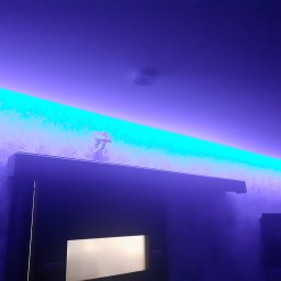 LUKA elektro - Nowoczesne oświetlenie LED w kolorze turkusowym, zamontowane pod sufitem, tworzące dekoracyjny efekt świetlny na ścianie z wzorzystą tapetą w odcieniach fioletu.