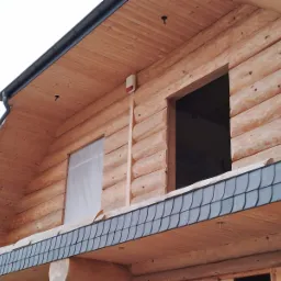 Zewnętrzny widok drewnianego domu z bali z zainstalowanym alarmem na ścianie, okno zabezpieczone folią ochronną podczas prac wykończeniowych.