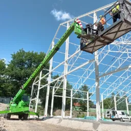 Montaż konstrukcji stalowej hali z użyciem podnośnika koszowego, dwóch pracowników w kamizelkach odblaskowych na wysokości, błękitne niebo w tle.