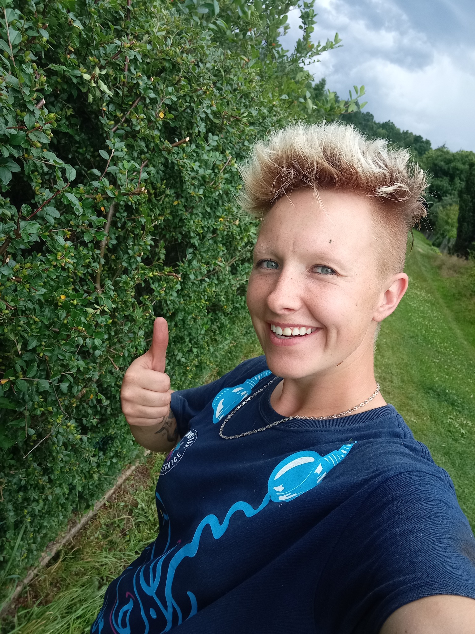 Uśmiechnięta kobieta z krótkimi blond włosami, w ciemnej koszulce z niebieskim nadrukiem, kciukiem w górę na tle przyciętego żywopłotu i trawnika.