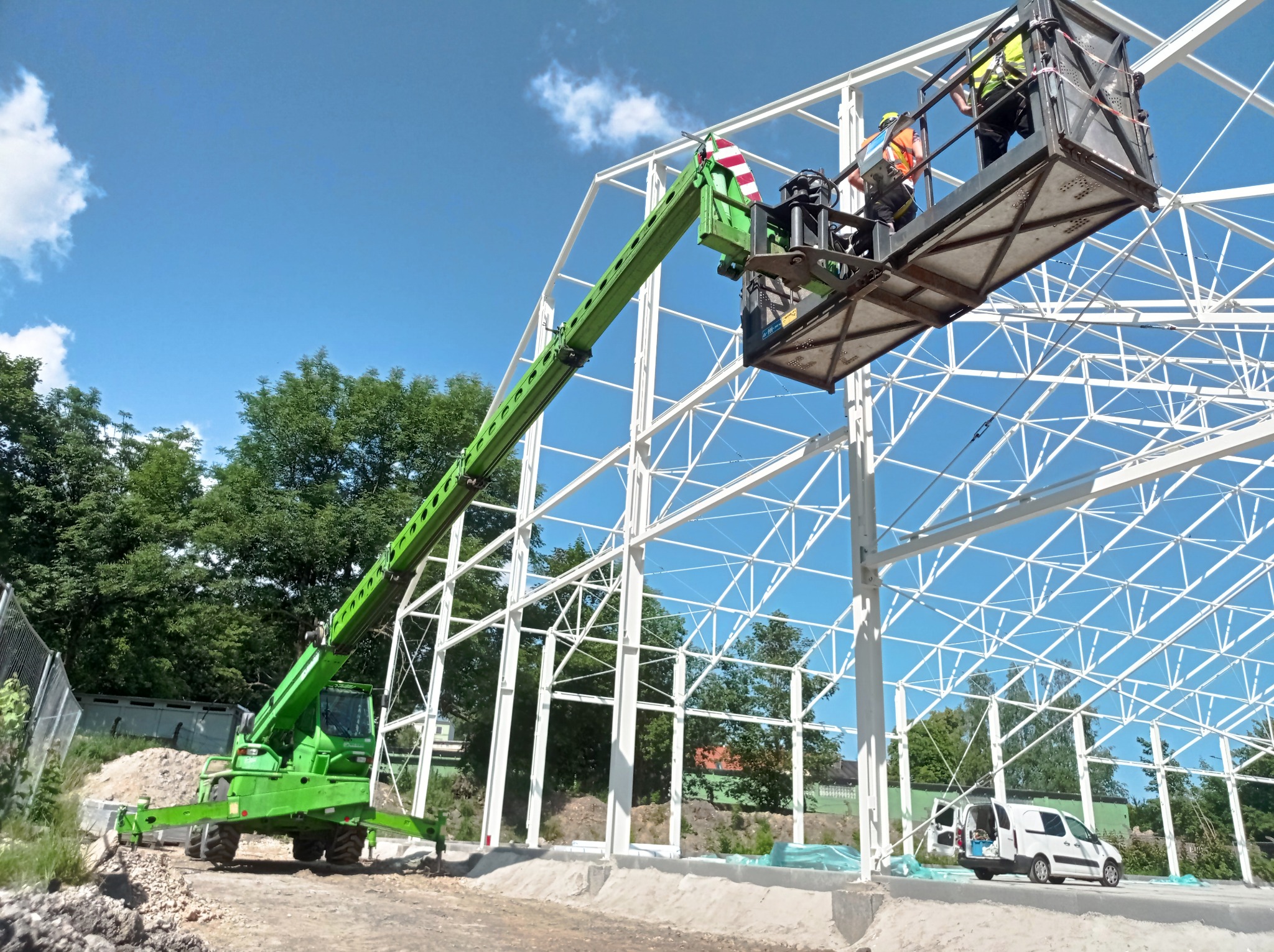 Montaż konstrukcji stalowej hali z użyciem podnośnika koszowego, dwóch pracowników w kamizelkach odblaskowych na wysokości, błękitne niebo w tle.