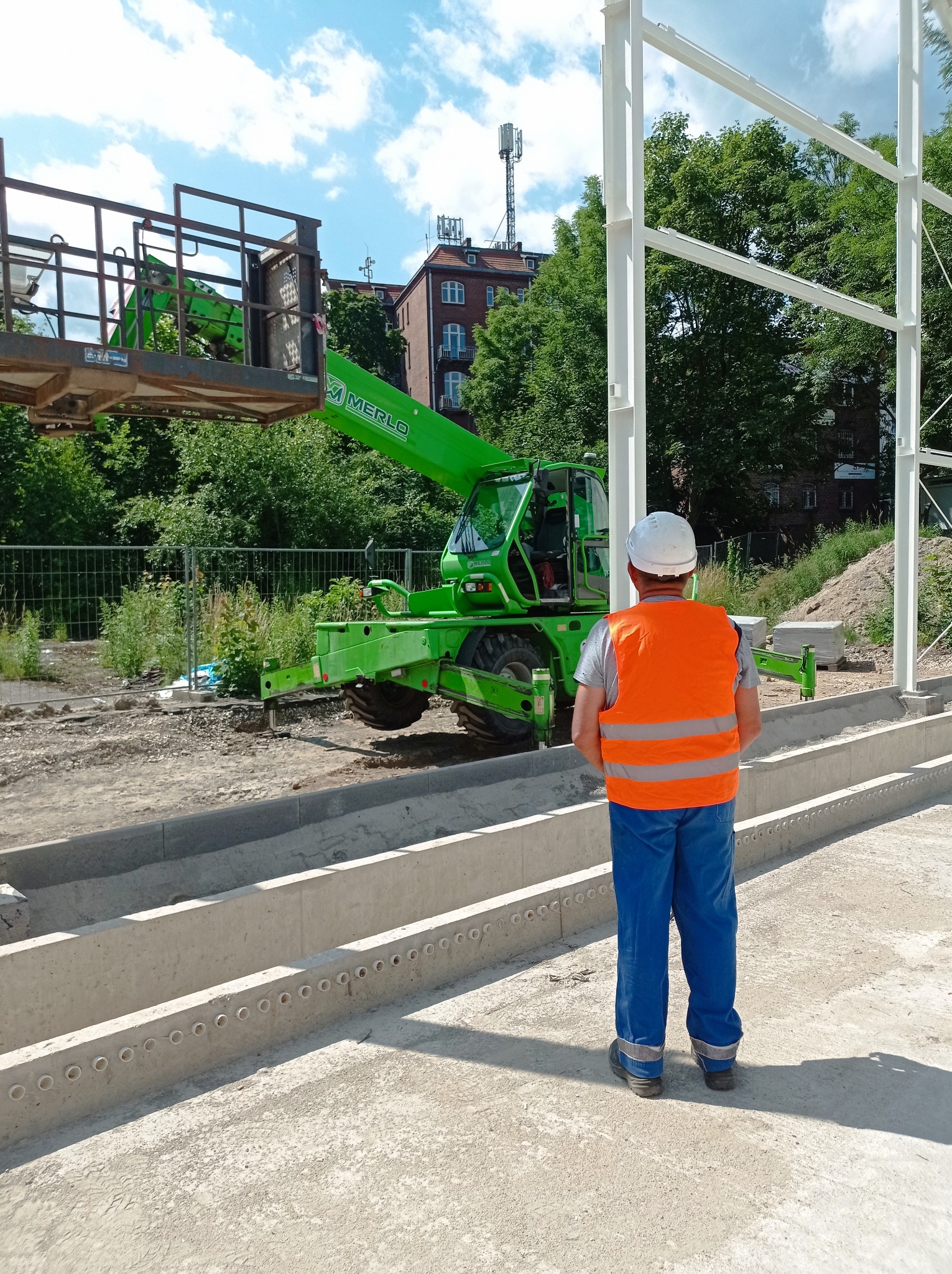 Pracownik w kasku i kamizelce odblaskowej stoi na placu budowy, obserwując zielony podnośnik teleskopowy Merlo i konstrukcję stalową budynku w tle.