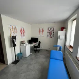 Gabinet lekarski w Krakowie z niebieskim kozetką, biurkiem z komputerem, plakaty anatomiczne na ścianach i dozownik do dezynfekcji przy wejściu.