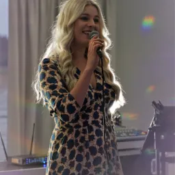 Uśmiechnięta blondynka w kwiecistej sukience śpiewa do mikrofonu na tle sprzętu nagłośnieniowego i świateł dyskotekowych.