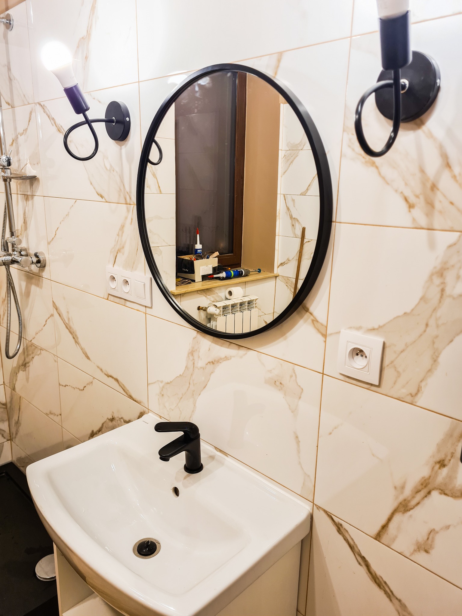 Wykończona łazienka z umywalką, armaturą w kolorze czarnym, okrągłym lustrem i lampami ściennymi o nietypowym kształcie, widoczne narzędzia na parapecie w odbiciu lustra.