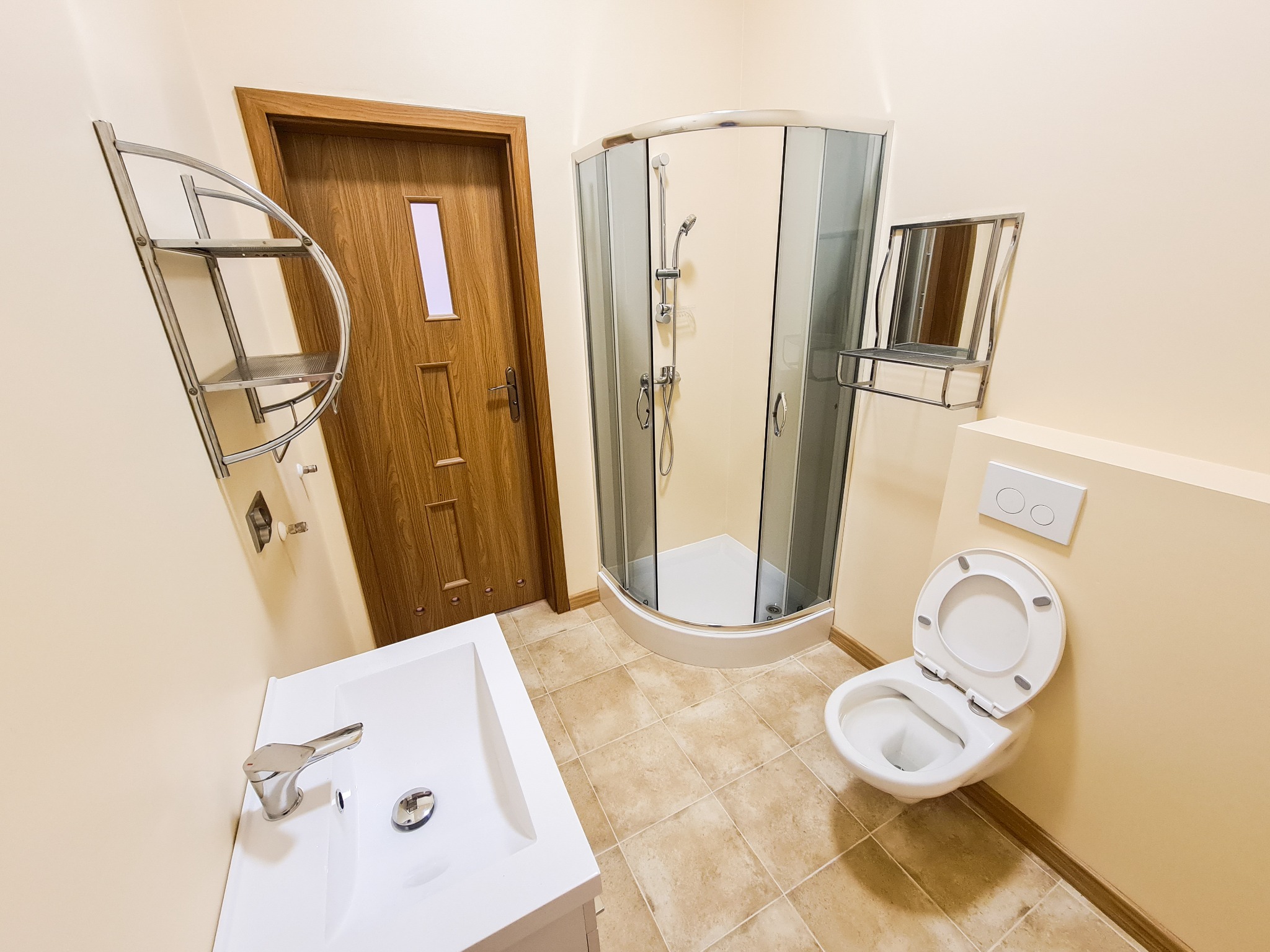 Kompletna łazienka po remoncie z kabiną prysznicową, umywalką, toaletą i drewnianymi drzwiami z małym oknem, wykończona w jasnych kolorach z beżowymi płytkami podłogowymi.