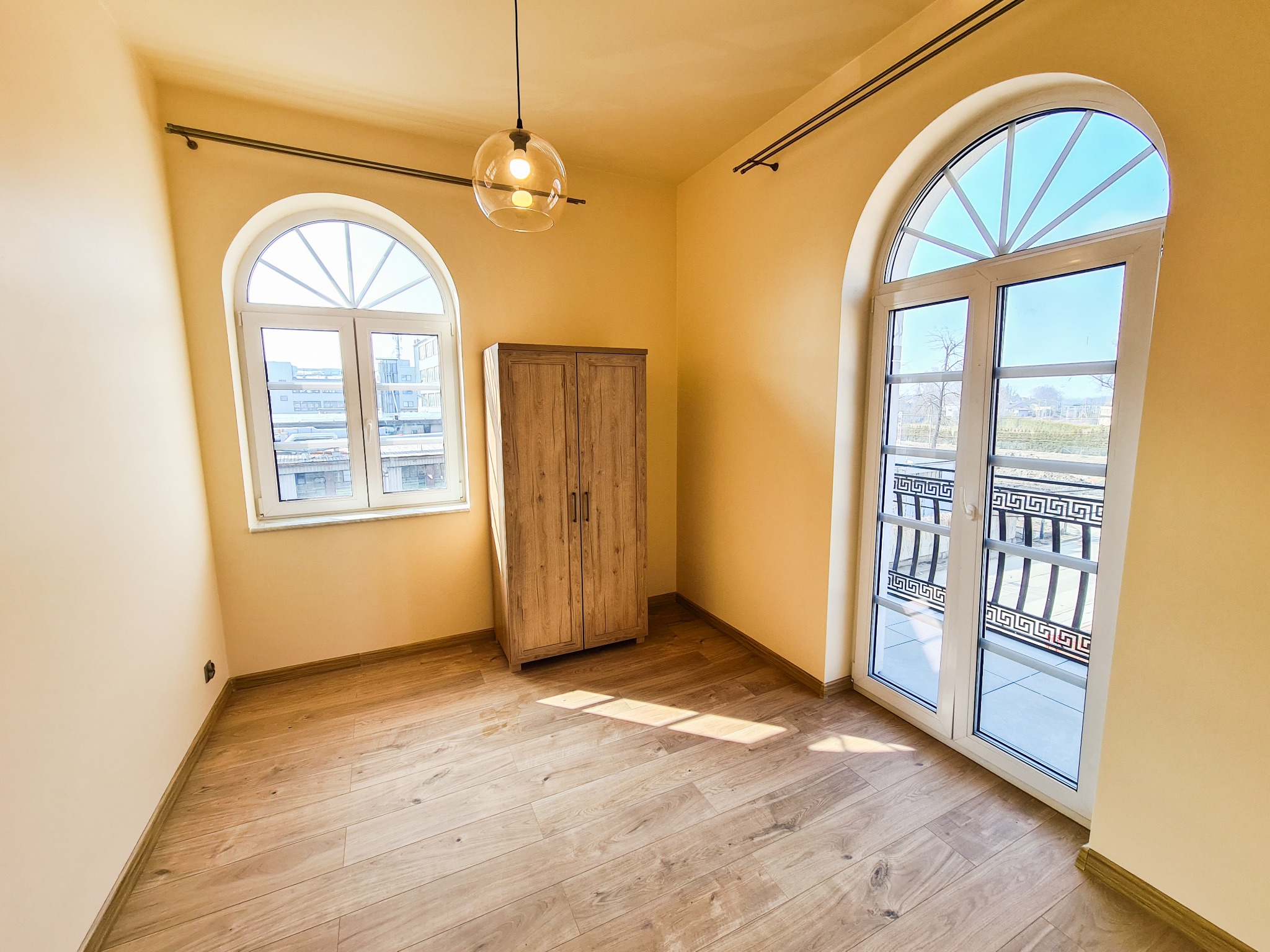 Jasne wnętrze z drewnianą podłogą, szafką z naturalnego drewna i dwoma łukowymi oknami, jedno z wyjściem na balkon z ozdobną balustradą, oświetlone nowoczesną lampą wiszącą.