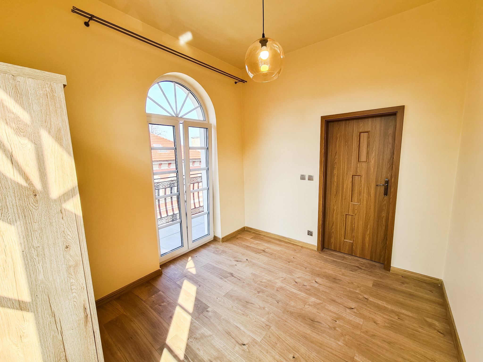 Pusty pokój z drewnianą podłogą, żółtymi ścianami, zaokrąglonym oknem balkonowym z balustradą na zewnątrz, drewnianymi drzwiami i wiszącą szklaną lampą. Na lewo częściowo widoczna szafa z jasnego...