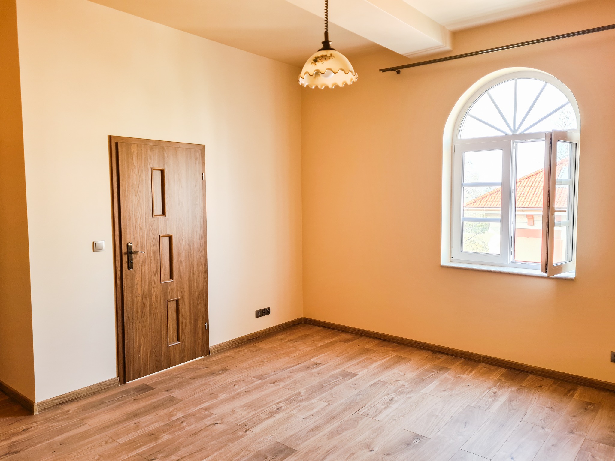 Pusty pokój po remoncie z drewnianą podłogą, drzwiami z prostokątnymi wstawkami i łukowym oknem z otwartym skrzydłem, oświetlony wiszącą lampą z kwiatowym motywem.