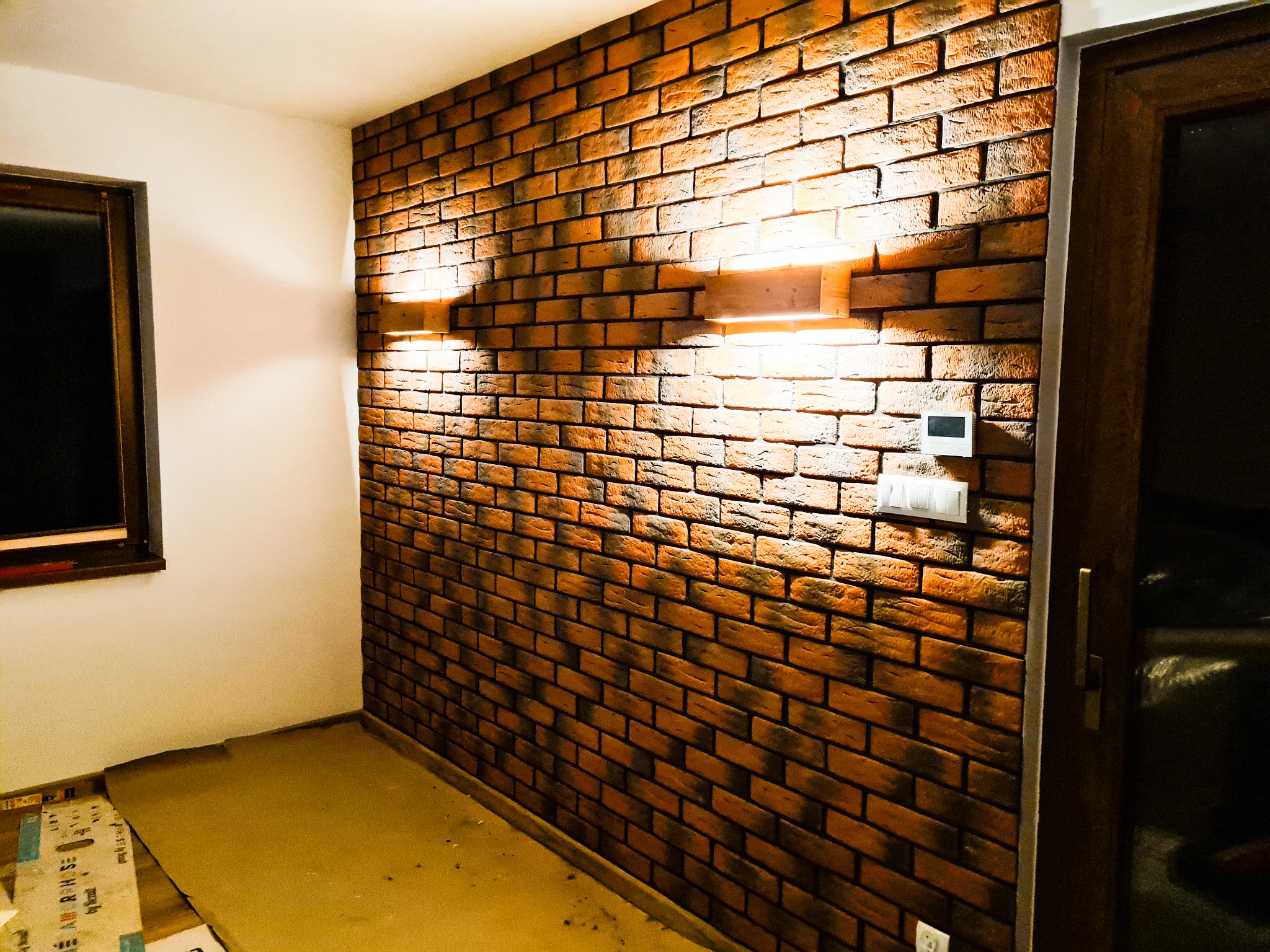 Ściana wykończona dekoracyjną cegłą klinkierową w odcieniach brązu i pomarańczy, oświetlona dwoma kinkietami, obok okno z ciemną ramą i drzwi, podłoga zabezpieczona kartonem.