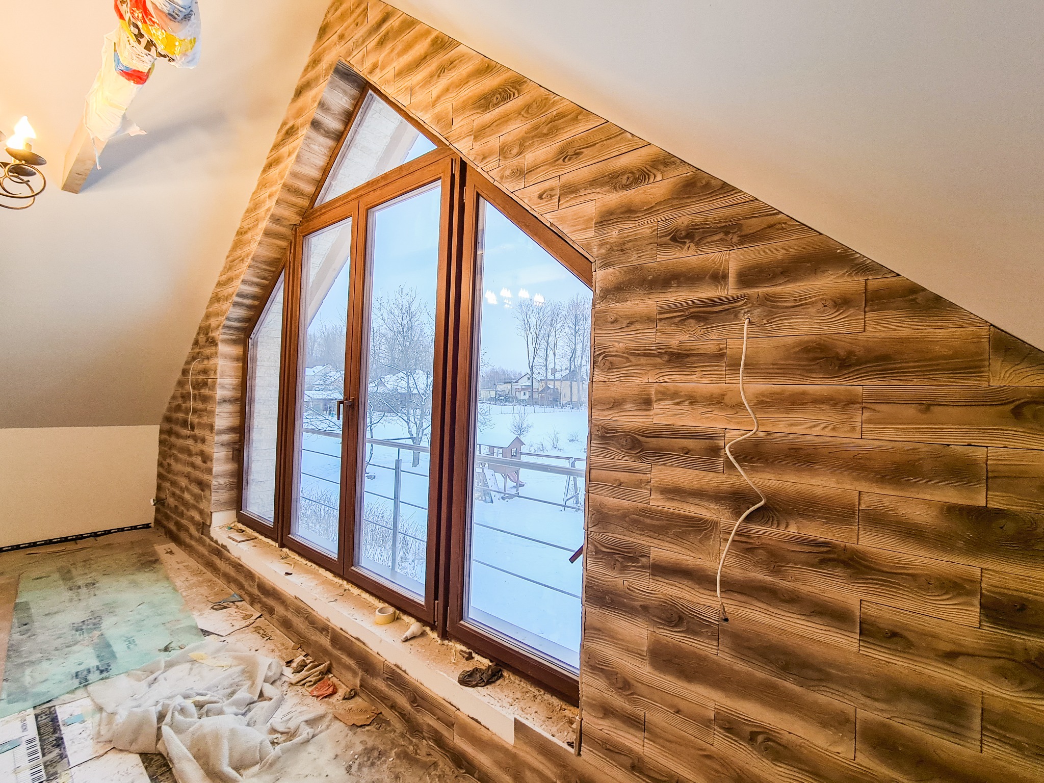 Poddasze z oknem w drewnianej ramie i boazerią imitującą drewno na ścianach, widoczne ślady prac wykończeniowych na podłodze.
