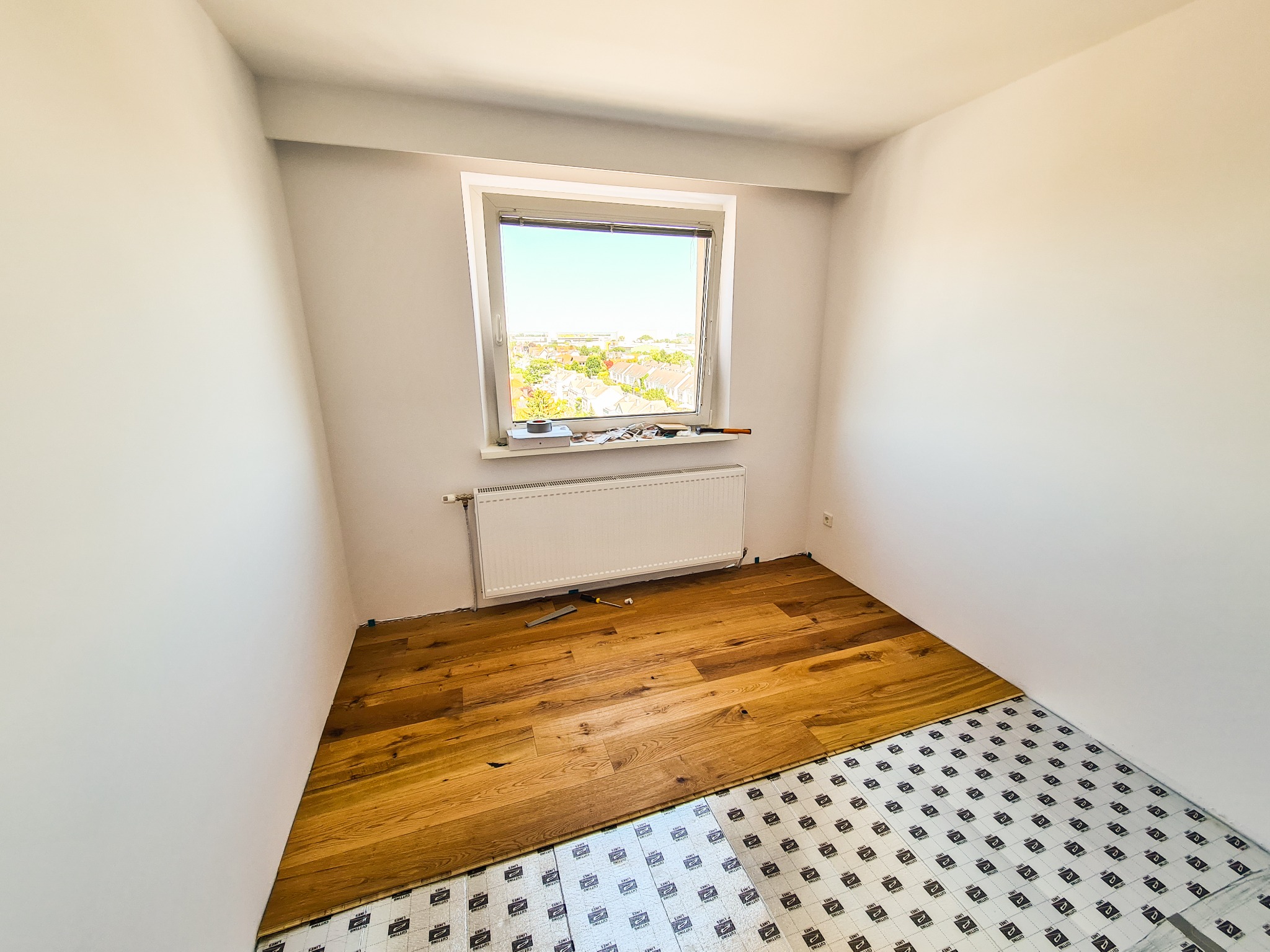 Układanie drewnianej podłogi w pokoju z widokiem na miasto przez okno, widoczna warstwa izolacyjna pod panelami.