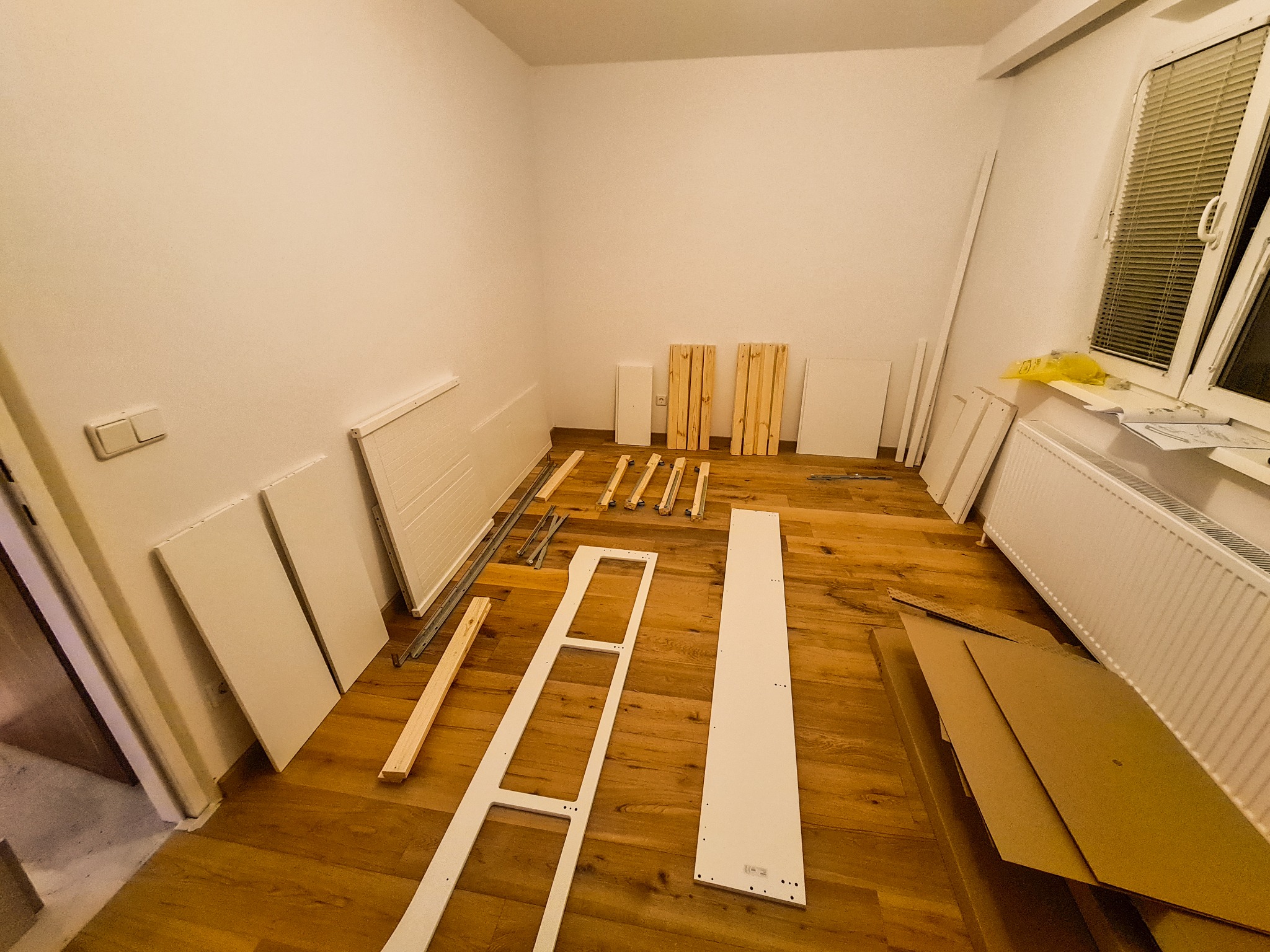 Rozpakowane elementy mebli, w tym białe panele, drewniane listwy i metalowe prowadnice, rozłożone na drewnianej podłodze w pustym pokoju podczas montażu.