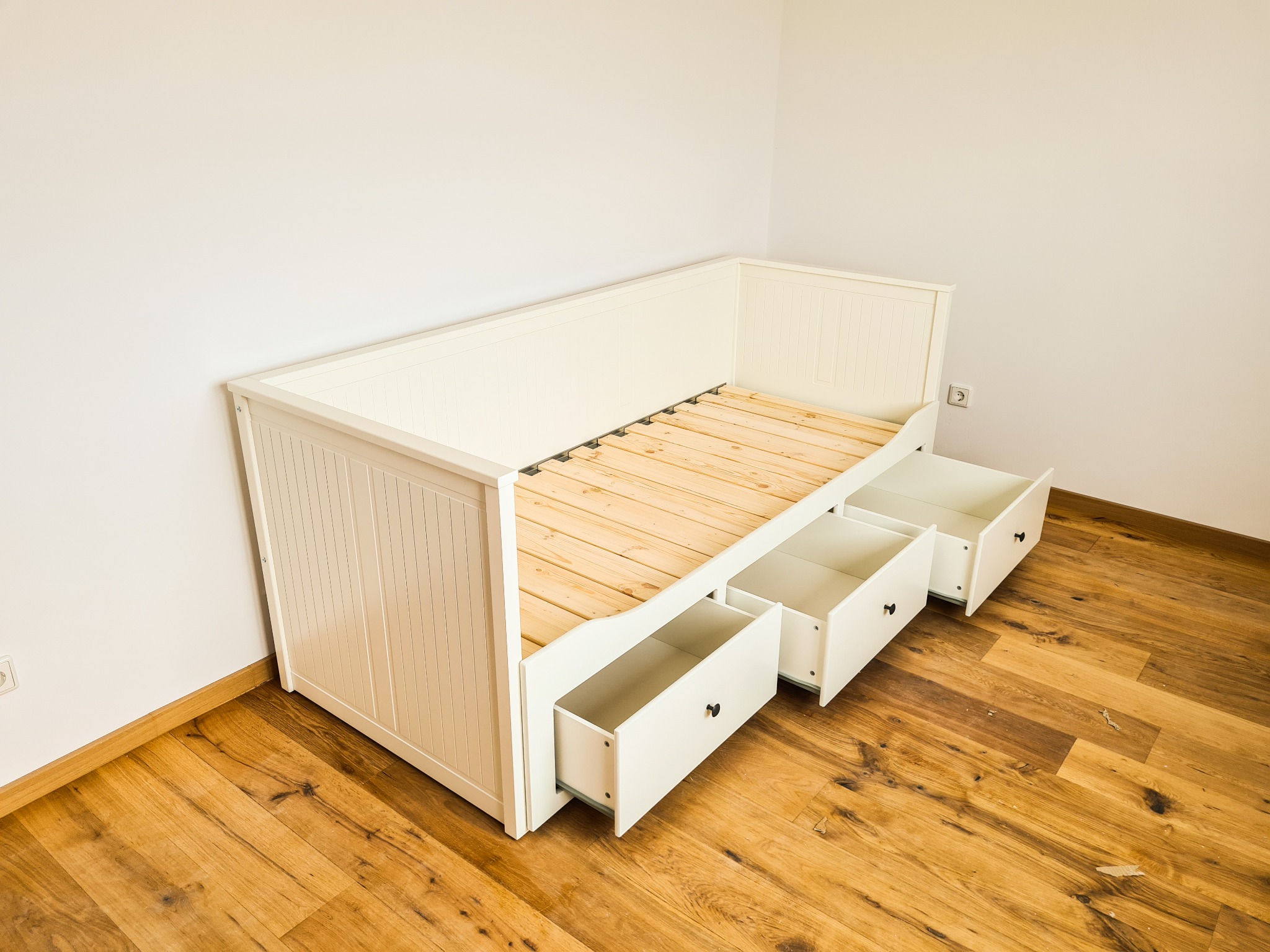 Biała, drewniana sofa z wysuwanymi szufladami na tle parkietu i białej ściany