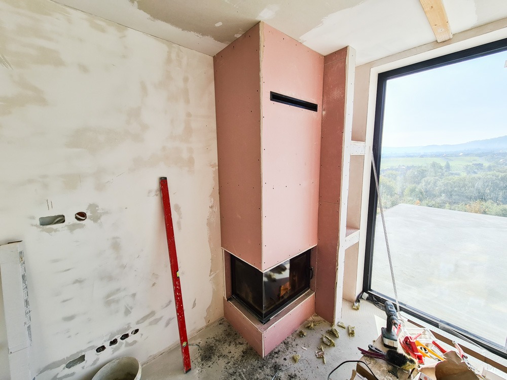 Wnętrze w trakcie prac: obudowa kominka z płyt kartonowo-gipsowych, ściana z widocznymi śladami szpachlowania, poziomica oparta o ścianę, okno z widokiem na krajobraz.