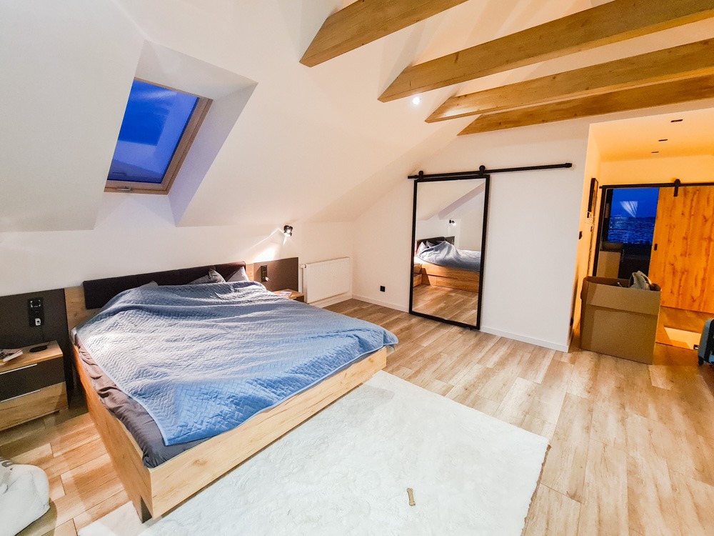 Sypialnia na poddaszu z drewnianymi belkami stropowymi, łóżkiem z jasnego drewna i przesuwnymi drzwiami do garderoby, widoczne odbicie łóżka w lustrze.