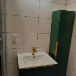 Nowoczesna łazienka z umywalką nablatową, złotą armaturą i szafką w odcieniach zieleni, oświetlona listwą LED.