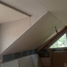 Montaż płyt kartonowo-gipsowych na poddaszu, widoczne mocowania i okno dachowe z widokiem na zieleń.