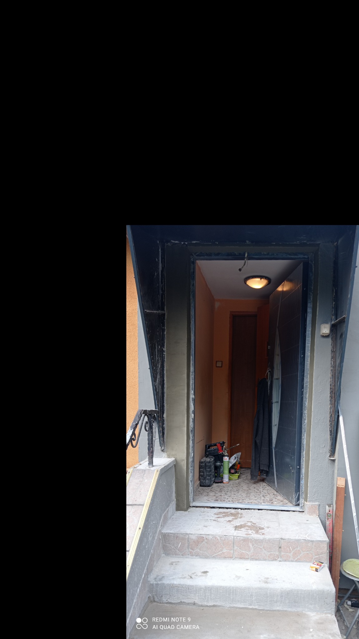 Schody wejściowe po nałożeniu szarej warstwy wykończeniowej, widoczne narzędzia i materiały budowlane w tle, drzwi wejściowe otwarte.