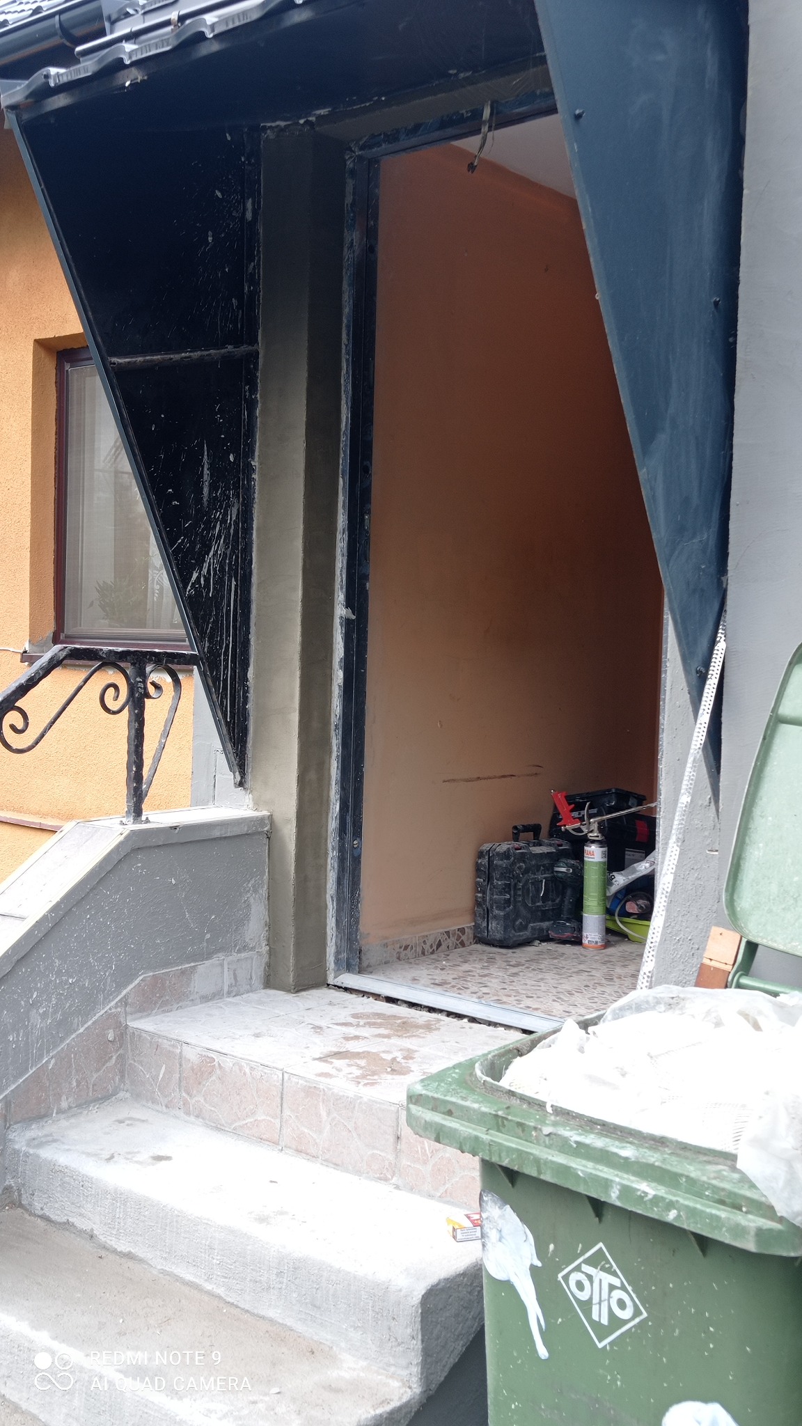 Prace murarskie przy wejściu do budynku: obrobione ościeże drzwi wejściowych, widoczne narzędzia i pojemnik na odpady budowlane.