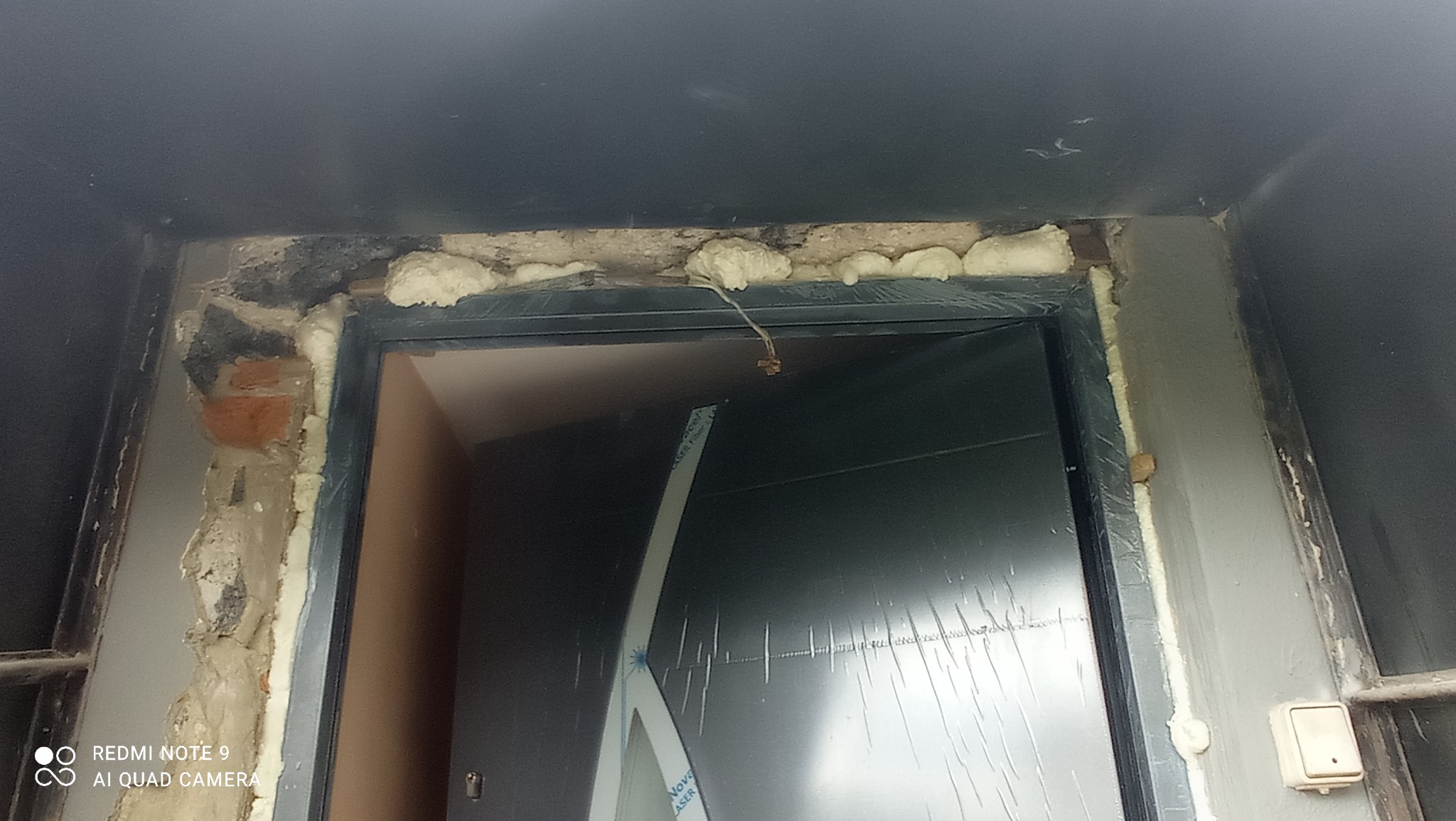 Montaż ościeżnicy drzwiowej z widoczną pianką montażową i fragmentami starego muru, zabezpieczona folią.