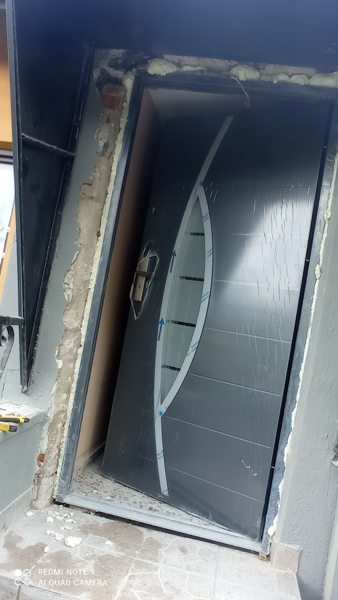 Montaż nowoczesnych, grafitowych drzwi wejściowych z owalnym przeszkleniem, widoczna piana montażowa wokół ościeżnicy i częściowo wykończone obramowanie.