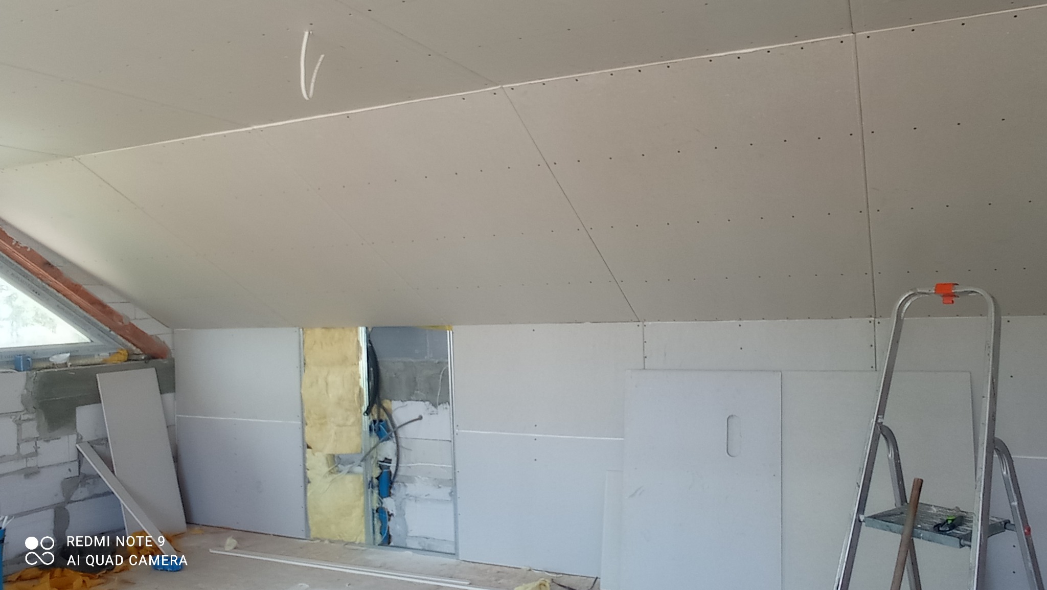 Poddasze w trakcie wykańczania, widoczne płyty gipsowo-kartonowe na ścianach i suficie, częściowo zakryta izolacja z wełny mineralnej, instalacja elektryczna w peszlach, drabina aluminiowa po...