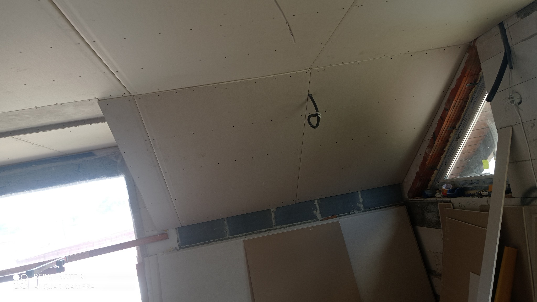 Montaż płyt gipsowo-kartonowych na poddaszu, widoczne śruby mocujące, okno dachowe i fragment instalacji elektrycznej.