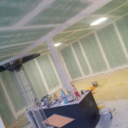 Poddasze w trakcie ocieplania z widoczną konstrukcją sufitu podwieszanego z płyt gipsowo-kartonowych, narzędziami i materiałami budowlanymi na prowizorycznym stole oraz stojącym grzejnikiem gazowym.