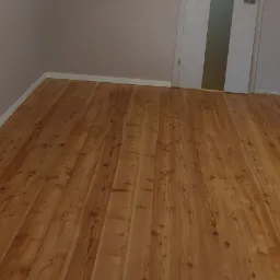 Wycyklinowana i polakierowana podłoga z desek sosnowych w pokoju z białymi listwami przypodłogowymi i drzwiami z mleczną szybą.