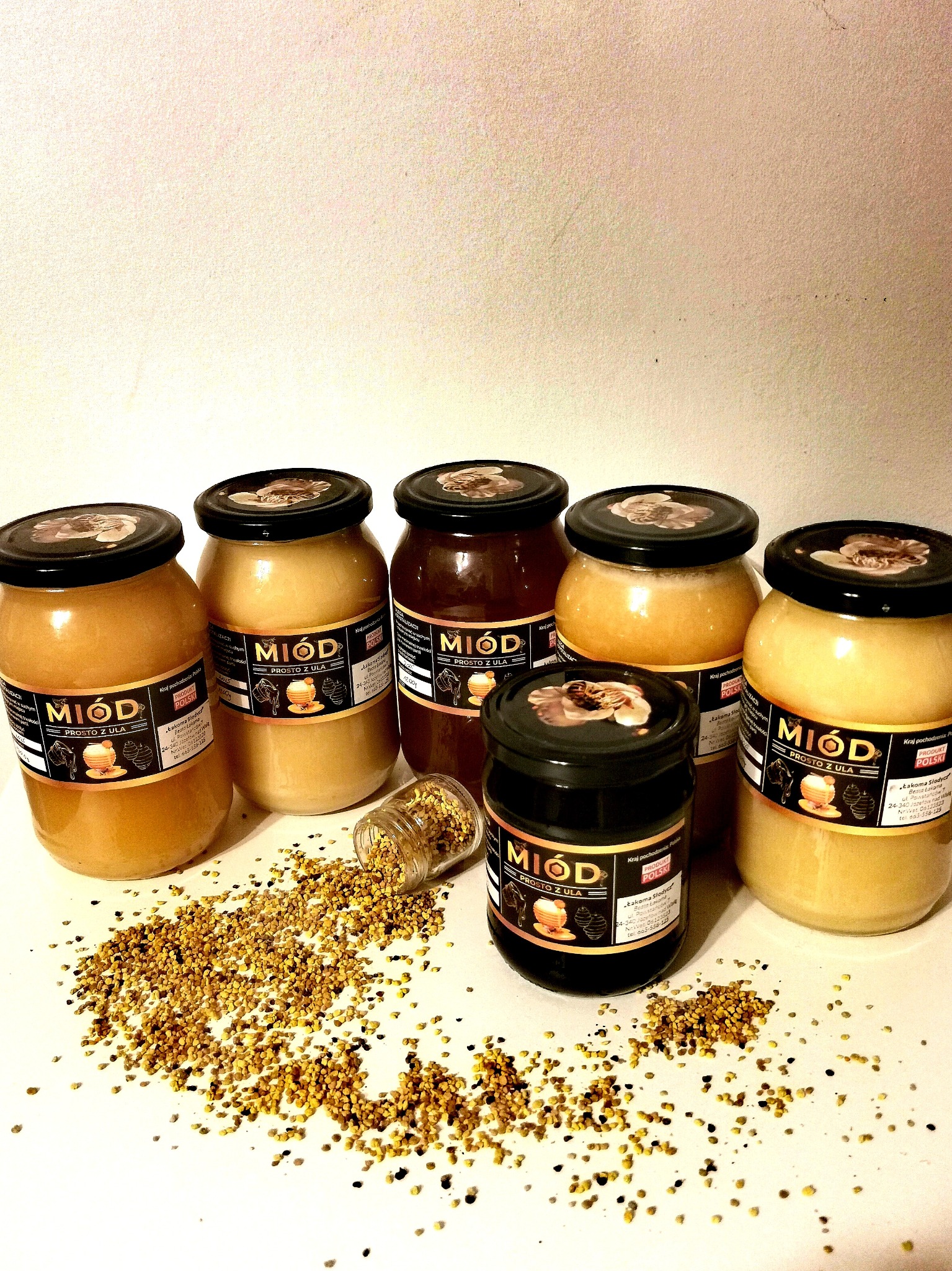 Sześć słoików z miodem o różnej barwie, od jasnego do ciemnego, ustawionych na białym tle, z rozsypanym pyłkiem kwiatowym i przewróconym małym słoiczkiem obok.