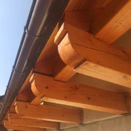 Widok z dołu na drewnianą konstrukcję okapu z brązowymi rynnami, widoczny fragment dachu domu w tle.