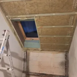 Montaż okna dachowego w trakcie ocieplania poddasza, widoczna warstwa izolacji paroprzepuszczalnej z nadrukiem FOLIAREX, aluminiowa drabina oparta o ścianę, fragment ściany z pustaków.