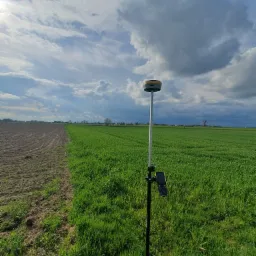 Statyw z odbiornikiem GNSS na pograniczu zaoranego pola i zielonego pola uprawnego pod błękitnym, pochmurnym niebem.