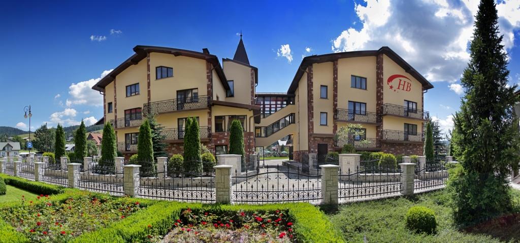 Hotel z beżową elewacją, balkonami z czarną balustradą, połączony przeszklonym łącznikiem, otoczony ozdobnym metalowym ogrodzeniem i zadbanym ogrodem z czerwonymi kwiatami.